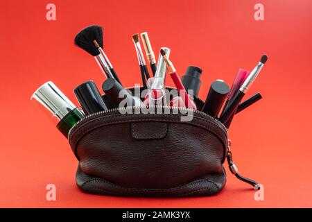 Make up cosmetics bag with professional accessories. Lipsticks, nail polish and brushes in a brown leather purse against red color background Stock Photo