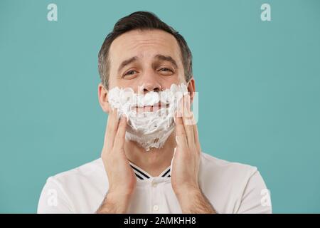 Head and shoulders portrait of smiling adult man shaving and looking at camera while enjoying morning routine standing against blue background, copy space Stock Photo