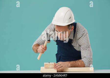 Portrait of frowning senior worker hammering nails while building wooden structure against blue background, copy space Stock Photo