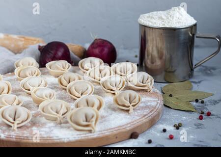 Uncooked, frozen dumplings on a wooden board. In the background is a metal mug with flour, next to pepper, greens and onions on a light background. Wi Stock Photo