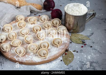 Uncooked, frozen dumplings on a wooden board. In the background is a metal mug with flour, next to pepper, greens and onions on a light background. Wi Stock Photo