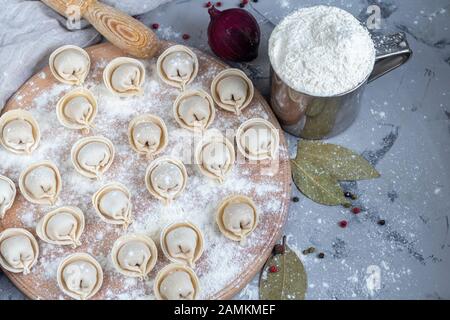Uncooked, frozen dumplings on a wooden board. In the background is a metal mug with flour, next to pepper, greens and onions on a light background. Wi Stock Photo