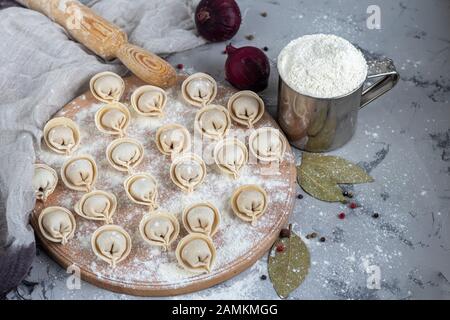 Uncooked, frozen dumplings on a wooden board. In the background is a metal mug with flour, next to pepper, greens and onions on a light background. Wi Stock Photo