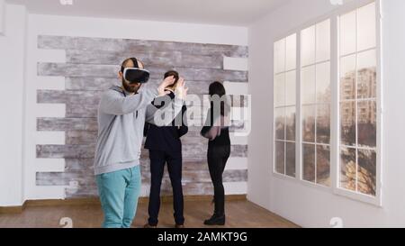 Caucasian man using virtual reality headset in empty apartment while a woman is talking with real estate agent. Stock Photo