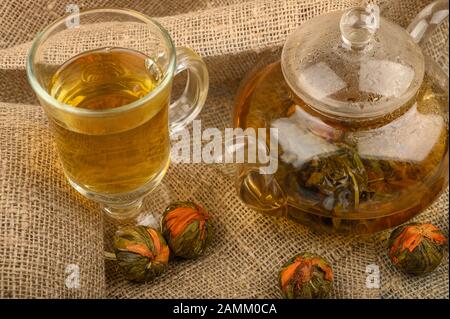 Flower tea brewed in a glass teapot, a glass of tea and balls of flower tea on a background of rough homespun fabric. Close up Stock Photo