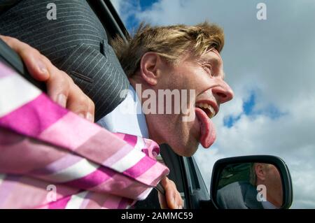 Face of an excited traveling businessman hanging out the car window with tongue sticking out and tie blowing in the wind Stock Photo