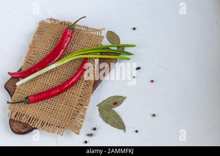 Red hot chili peppers on a wooden board. Near green onions, bay leaves, peppercorns. On white background. Copy space. Stock Photo