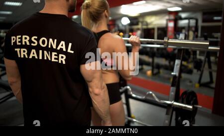 Young female doing weight bar lifting exercise under control of her trainer Stock Photo