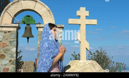 Religious woman in headscarf praying near cross, looking into sky with hope Stock Photo