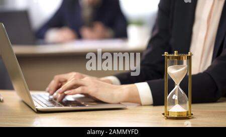 Female working on laptop at office, hourglass measuring time, efficiency at work Stock Photo