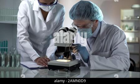 Medical researcher looking through microscope and colleague taking notes, lab Stock Photo