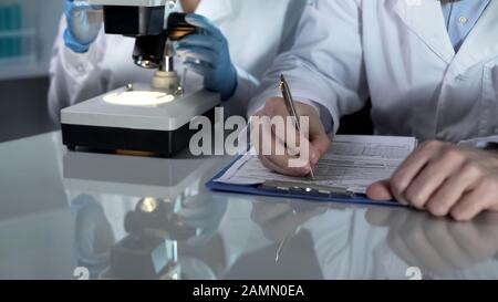 Lab worker filling paper forms, his assistant viewing samples under microscope Stock Photo