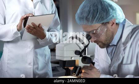 Researcher working with microscope, assistant recording comments on tablet Stock Photo