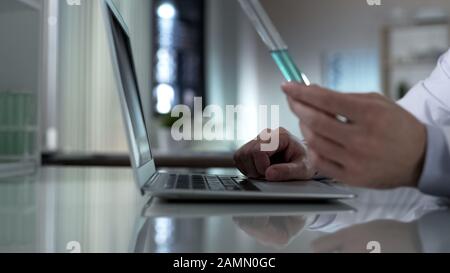 Biochemist holding tube with turquoise liquid, typing test results on laptop Stock Photo