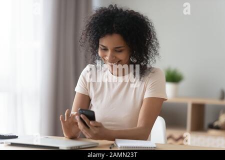 Positive african woman sit at desk typing message using smartphone Stock Photo