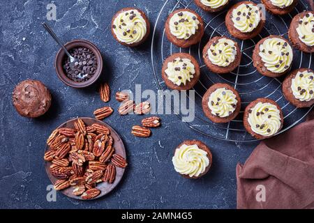 Chocolate pecan cupcakes topped with buttercream swirl icing on a metal cake rack on a concrete table with ingredients, horizontal view from above, fl Stock Photo