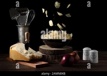 Potato chips falling in a bowl on a table with parmesan cheese, onions, salt and pepper - Dark mood Stock Photo