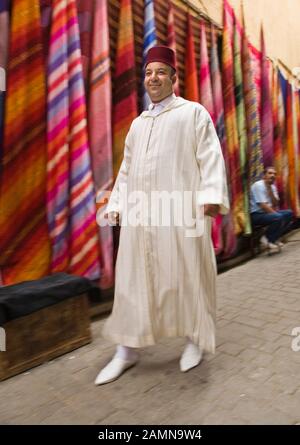 Man in traditional Moroccan clothing walking past colourful fabrics for sale in the Souk, Medina, Fez (Fes), Morocco, North Africa, Africa Stock Photo