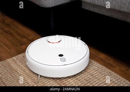 Robotic vacuum cleaner runs under sofa in room on laminate floor modern smart cleaning technology housekeeping. Stock Photo