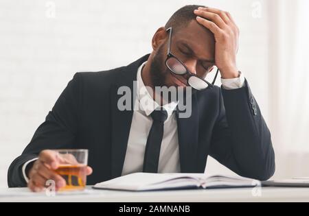 Exhausted afro man in suit drinking alcohol, touching his head Stock Photo