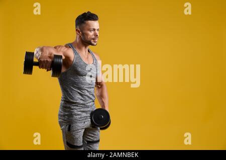 Side view of muscular bodybuilder doing exercise with dumbbells. Good looking athletic gentleman lifting weight. Isolated on yellow studio background with copy space. Concept of bodybuilding. Stock Photo