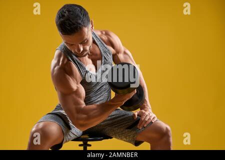 Side view of handsome bodybuilder sitting on stool and lifting heavy weight. Strong young man pumping iron. Isolated on yellow studio background. Concept of bodybuilding and sport. Stock Photo