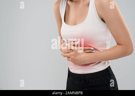 Clolse up of bending incognito caucasian woman in casual suit posing over gray isolated background and touching top of stomach because of ache. Side crop of brunette suffering from abdominal pain. Stock Photo