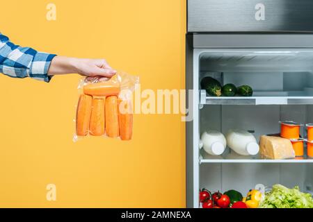cropped view of woman holding carrots in vacuum package near open fridge with fresh food on shelves isolated on yellow Stock Photo