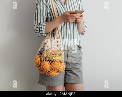 Unrecognizable young woman using smartphone standing with mesh bag on shoulder. Stylish young woman standing with mesh shopping bag on shoulder near light wall and using smartphone. Stock Photo