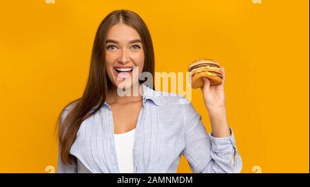 Cheerful Lady Holding Burger Standing On Yellow Studio Background, Panorama Stock Photo