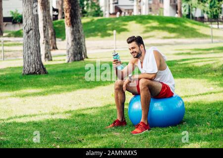 happy bearded man looking at sport bottle while sitting on fitness ball Stock Photo