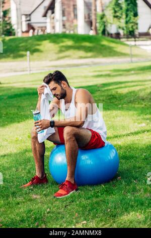 bearded man holding sport bottle while sitting on fitness ball and wiping sweat Stock Photo
