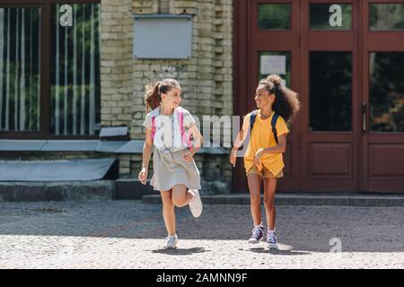 two cheerful multiethnic schoolgirls smiling while running in schoolyard Stock Photo