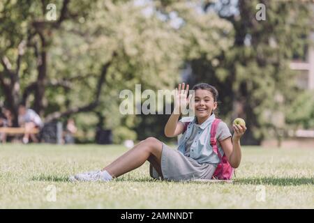 cheerful schoolgirl waving hand while sitting on lawn in park and looking at camera Stock Photo