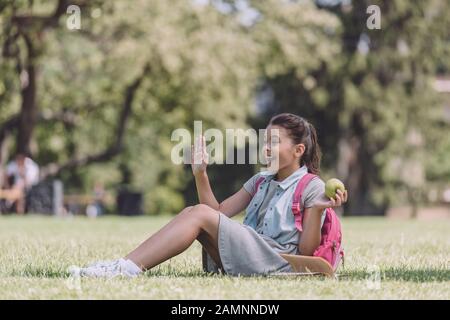 cheerful schoolgirl holding apple and waving hand while sitting on lawn in park Stock Photo