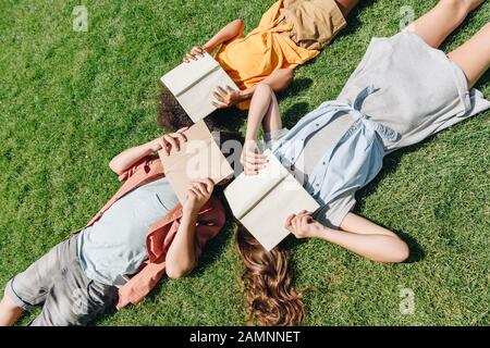 top view of three multicultural schoolkids lying on lawn and covering faces with books Stock Photo