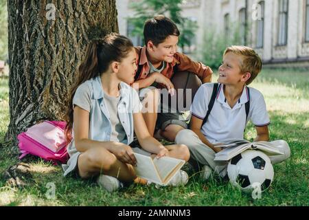 three cheerful schoolkids talking while sitting on lawn under tree