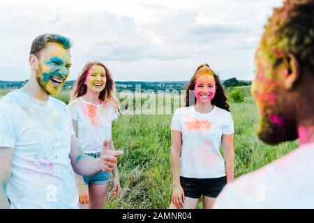 selective focus of happy man with holi paints on face pointing with finger at african american friend near girls Stock Photo
