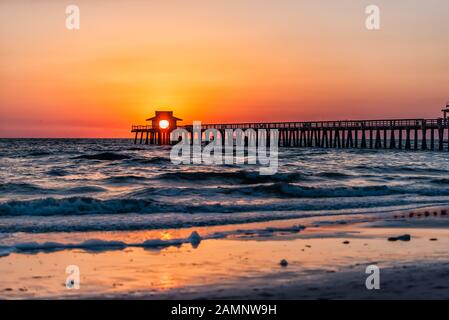 Naples, Florida colorful red sunset in gulf of Mexico with sun path reflection and Pier wooden jetty silhouette with horizon and ocean waves Stock Photo