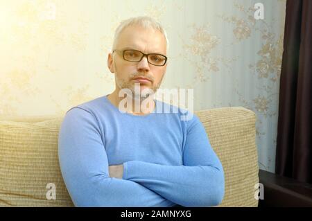 A handsome young man in a blue sweater and glasses sits and looks at the camera. Close-up Stock Photo