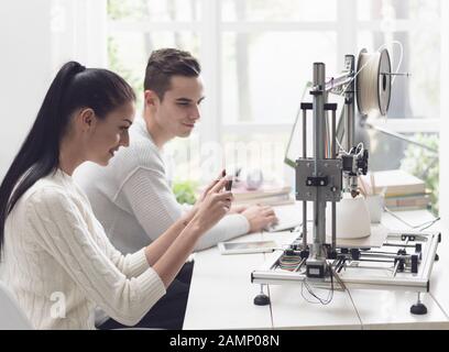 Engineering students printing prototype models using a 3D printer, the girl is taking pictures with a smartphone and sharing online Stock Photo