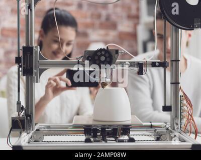 Engineering students printing prototype models using a 3D printer, the girl is taking pictures with a smartphone and sharing online Stock Photo