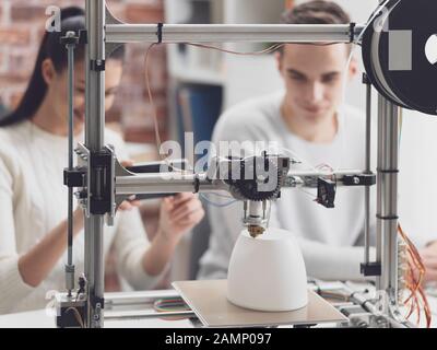 Engineering students printing prototype models using a 3D printer, the girl is taking pictures with a smartphone and sharing online Stock Photo