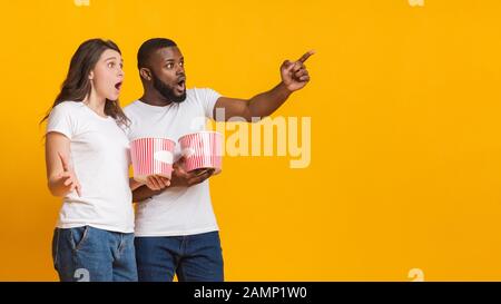 Surprised interracial friends holding popcorn buckets and pointing at copy space Stock Photo