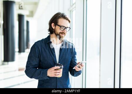 Smiling businessman is holding in hand cup of coffee and having pleasant cellphone conversation during work break, while is standing in office. Stock Photo