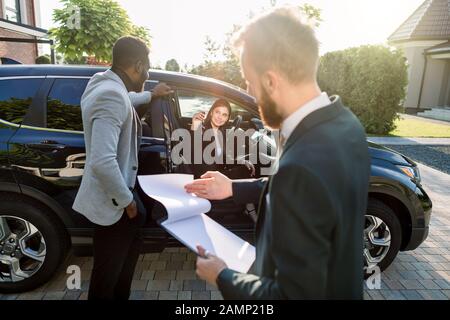 Happy multiethnical couple, African man and Caucasian woman, buying the car, black crossover, woman is sitting in the car and holding car keys. Young Stock Photo