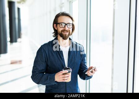 Smiling businessman is holding in hand cup of coffee and having pleasant cellphone conversation during work break, while is standing in office. Stock Photo