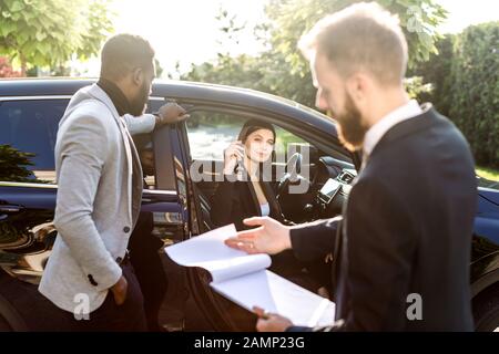 Happy multiethnical couple, African man and Caucasian woman, buying the car, black crossover, woman is sitting in the car and holding car keys. Young Stock Photo