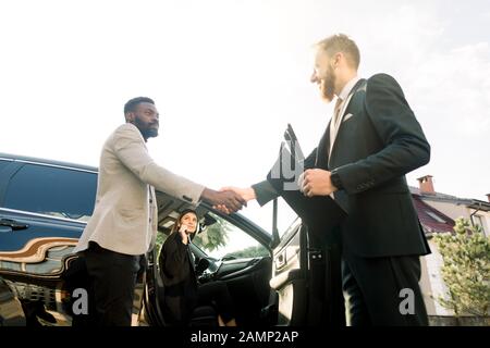 Sales situation in a car dealership, young Caucasian man dealer is shaking hands with a young African businessman, young woman is sitting in the car Stock Photo