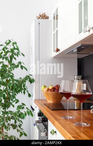 Two glasses of red wine stand on wooden tabletop of scandi kitchen with black wall on background of household appliances, cup with apples Stock Photo